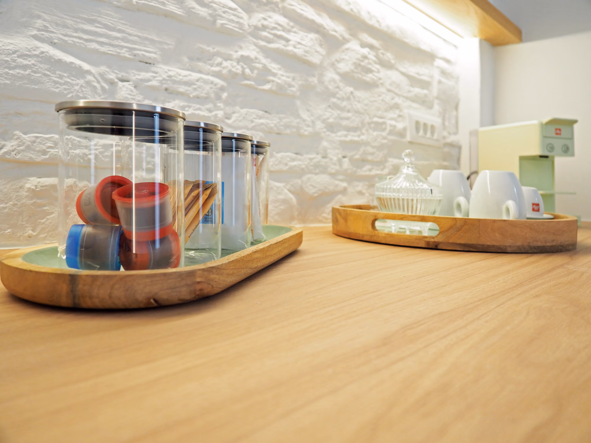 Wooden counter with trays holding jars, coffee pods, cups, and a coffee machine against a white brick wall.