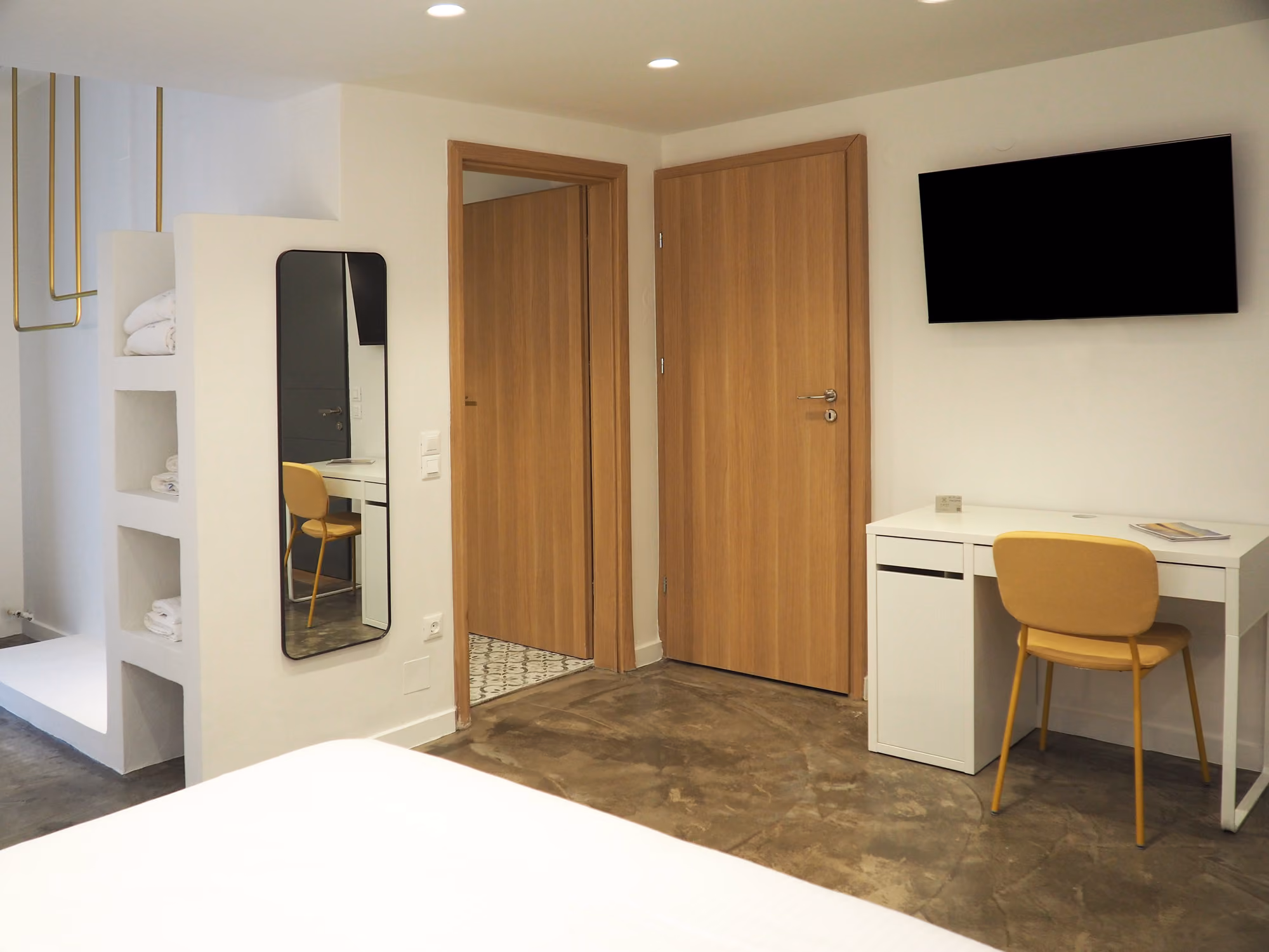 Modern hotel room corner with white desk, yellow chair, wall mirror, wooden doors, and mounted TV.